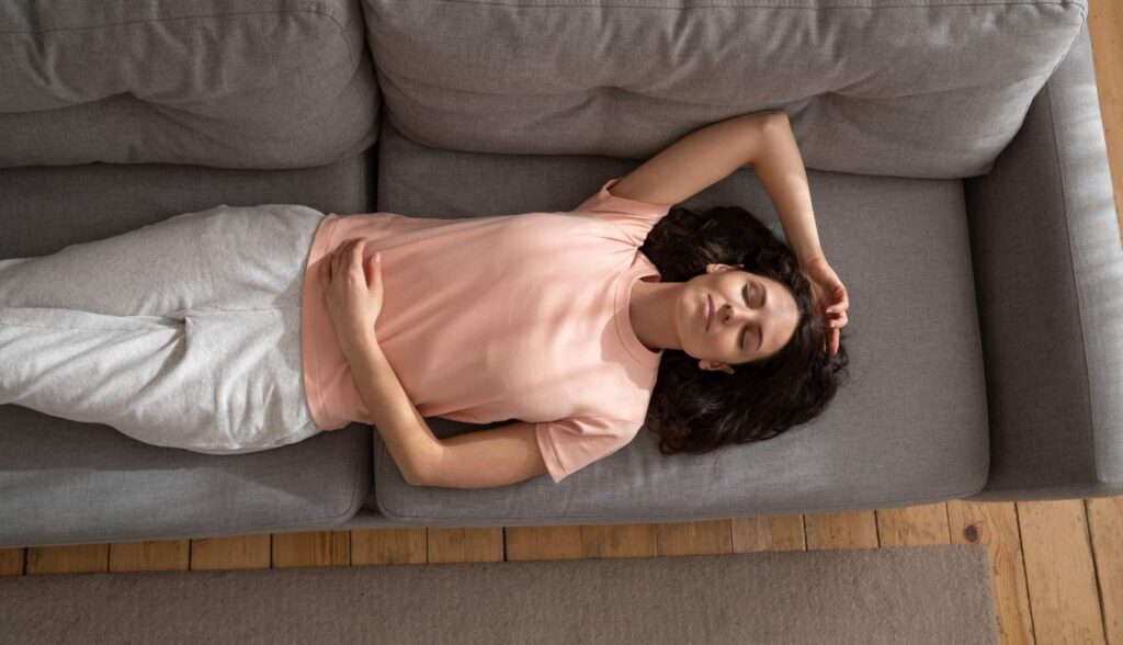 Young woman sleeping on couch with arm up at home, closing eyes and taking a break