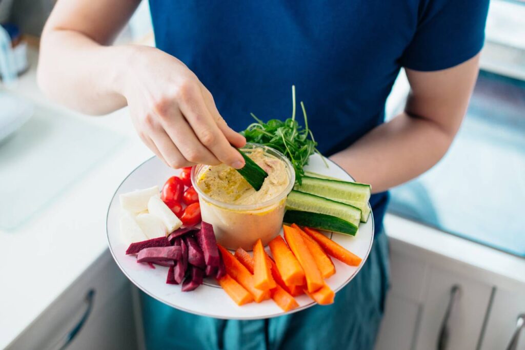 Healthy Snacking Tips - man dipping cucumber stick in hummus on the kitchen